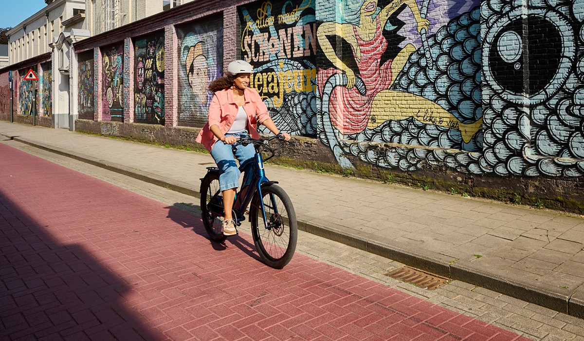 On the road in the city: Woman riding her fast Stromer ST1 e-bike in deep petrol along a painted wall. 